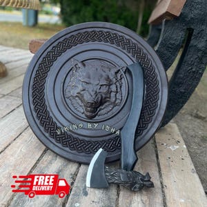 May include: A dark brown circular wooden shield featuring a carved wolf head in the center, surrounded by an intricate Celtic knot border and the text VIKING BY ISHA. A matching dark wooden axe with a metal blade rests against the shield.
