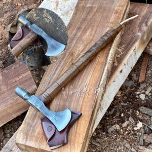 May include: A hand axe with a dark brown wooden handle featuring segmented ridges and a textured metal head. The axe includes a brown leather sheath with a snap button closure. The text Viking by isha is visible on the wooden handle and blade.