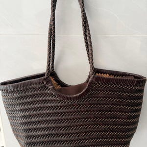 May include: A dark brown woven tote bag featuring a herringbone pattern throughout the body. The bag includes two long, braided shoulder straps and a solid brown fabric lining visible at the top opening, providing a structured yet flexible design.