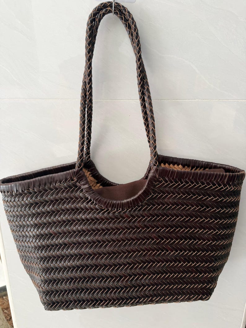 May include: A dark brown woven tote bag featuring a herringbone pattern throughout the body. The bag includes two long, braided shoulder straps and a solid brown fabric lining visible at the top opening, providing a structured yet flexible design.