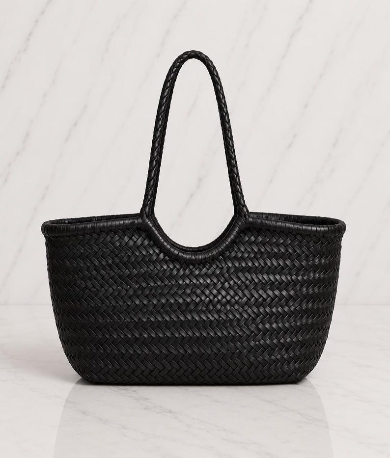 May include: A black tote bag featuring a woven construction with a consistent herringbone pattern throughout the body. The bag includes two integrated, braided handles that extend from the top edge, creating a rounded opening for carrying.