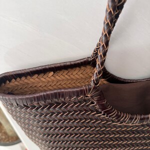 May include: A dark brown woven tote bag featuring a tight herringbone pattern throughout the exterior. The bag includes a braided handle and a solid dark brown fabric lining visible inside the top opening, showcasing detailed craftsmanship.