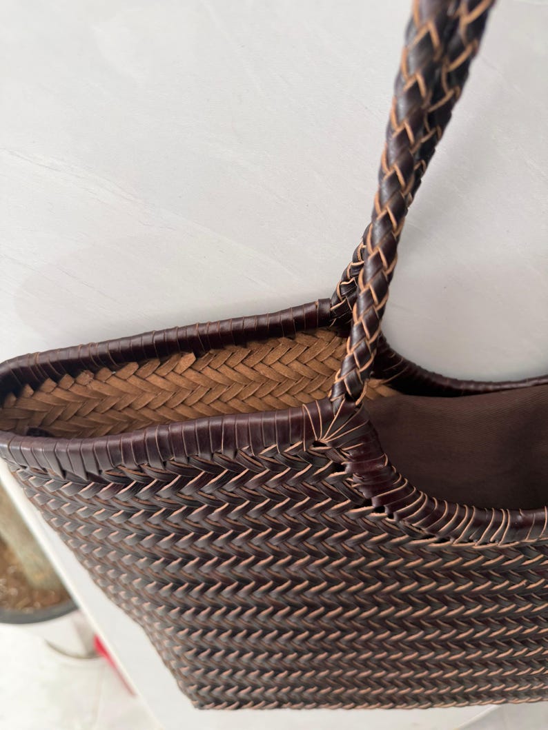 May include: A dark brown woven tote bag featuring a tight herringbone pattern throughout the exterior. The bag includes a braided handle and a solid dark brown fabric lining visible inside the top opening, showcasing detailed craftsmanship.