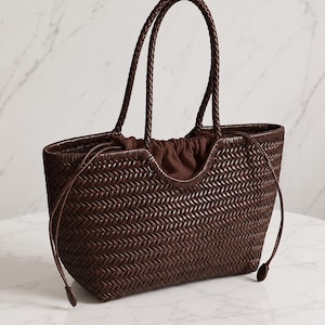 May include: A dark brown woven leather tote bag featuring a herringbone pattern, two braided top handles, and a drawstring closure with a matching brown fabric interior lining. The bag has a curved top opening and a structured rectangular base.