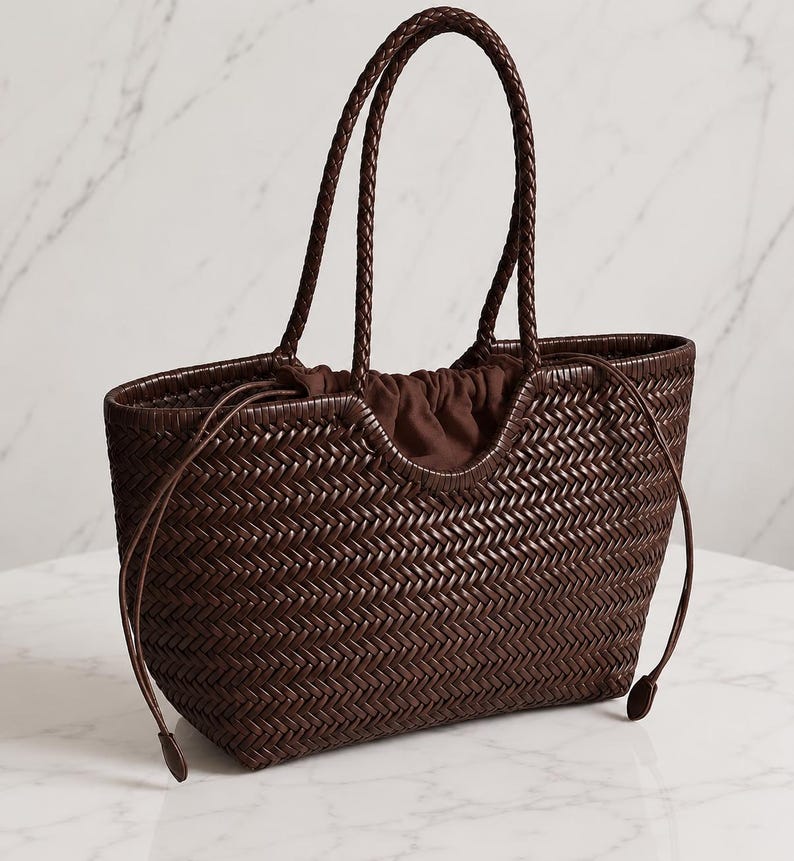 May include: A dark brown woven leather tote bag featuring a herringbone pattern, two braided top handles, and a drawstring closure with a matching brown fabric interior lining. The bag has a curved top opening and a structured rectangular base.