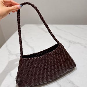 May include: A dark brown handbag featuring a woven leather construction with a rectangular body and a single braided shoulder strap. The bag has a consistent interlaced pattern throughout the exterior and the handle, creating a uniform texture.