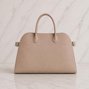 May include: A structured beige handbag with a curved top silhouette, two top handles, and side straps featuring silver-toned metal buckles. The bag has a smooth, solid-colored exterior with a horizontal seam detail across the upper section.