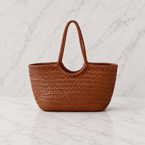 May include: A medium-sized, open-top tote bag crafted from woven brown leather. The bag features a consistent herringbone weave pattern throughout the body and two integrated, rounded leather handles that extend from the top rim.