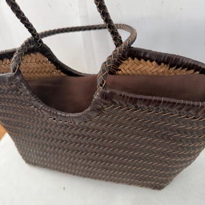 May include: A dark brown woven tote bag featuring a herringbone pattern with two braided handles. The interior is lined with a solid dark brown fabric, and the top edge of the bag is finished with a smooth, dark brown trim that matches the handles.