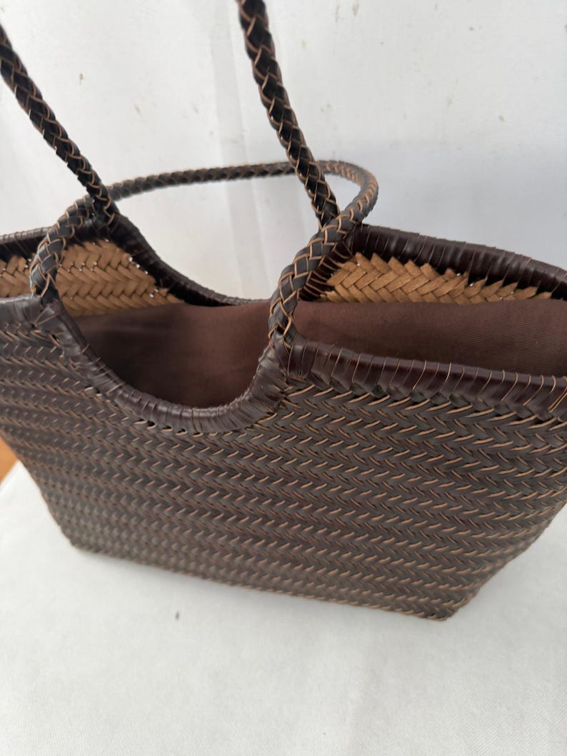 May include: A dark brown woven tote bag featuring a herringbone pattern with two braided handles. The interior is lined with a solid dark brown fabric, and the top edge of the bag is finished with a smooth, dark brown trim that matches the handles.