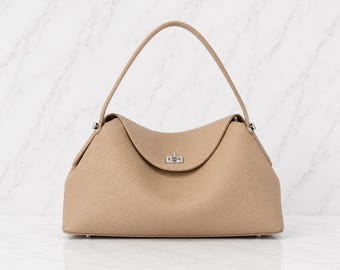May include: A beige leather handbag with a textured surface, featuring a single arched handle and a silver-toned metal turn-lock closure on the front flap. The bag has a structured base and a soft, curved silhouette with silver hardware accents.
