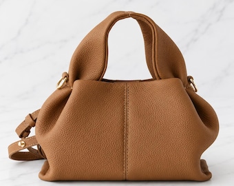May include: A tan handbag with a pebbled texture, featuring two rounded top handles, a central vertical seam, and gold-toned metal hardware. The bag includes a detachable, adjustable shoulder strap attached to side rings, with a soft, slouchy structure.