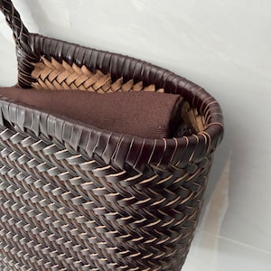 May include: A dark brown woven basket bag featuring a tight, intricate herringbone pattern. The rim is finished with a smooth, dark brown border, and the interior contains a solid dark brown fabric lining that extends slightly above the top edge.
