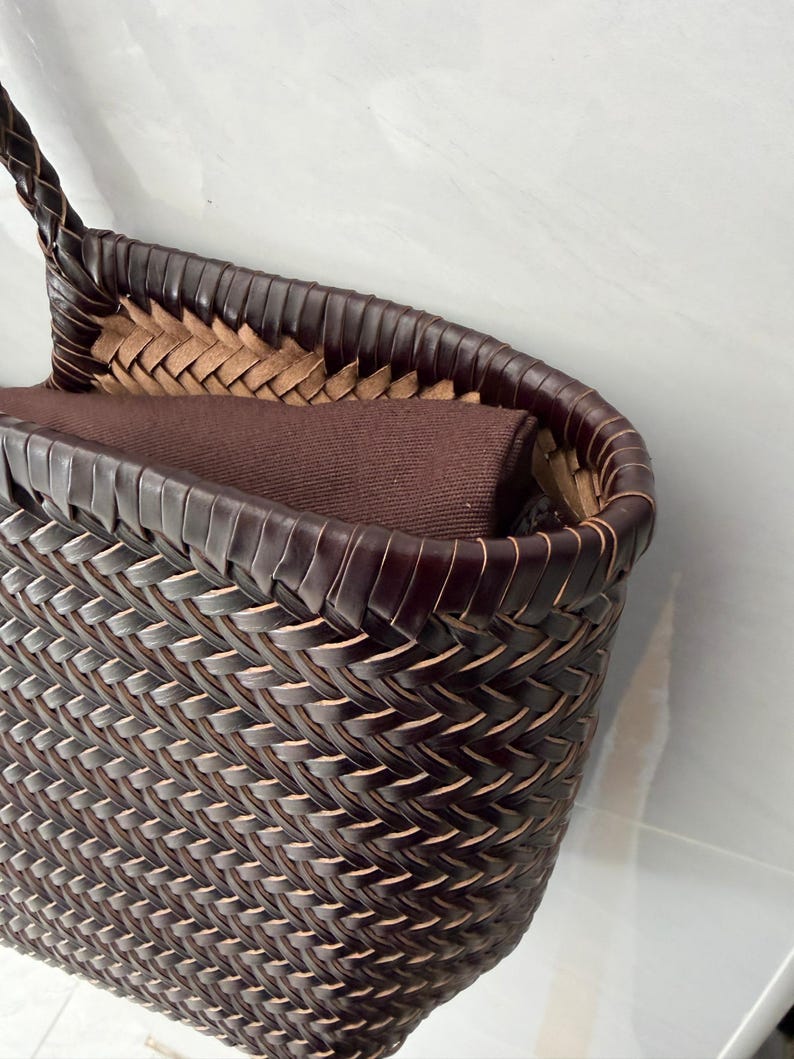 May include: A dark brown woven basket bag featuring a tight, intricate herringbone pattern. The rim is finished with a smooth, dark brown border, and the interior contains a solid dark brown fabric lining that extends slightly above the top edge.