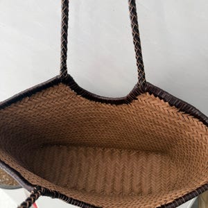 May include: A woven tote bag featuring a light brown herringbone pattern throughout the interior and exterior body. The bag includes a dark brown braided trim along the top opening and two matching dark brown braided shoulder straps.
