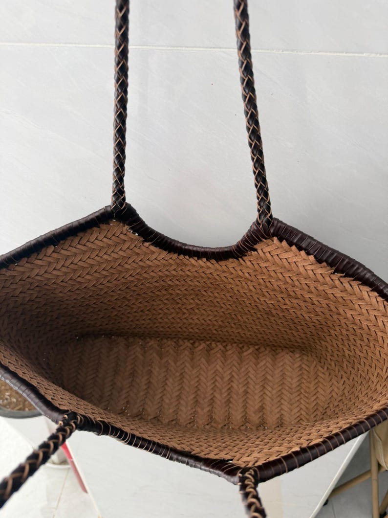 May include: A woven tote bag featuring a light brown herringbone pattern throughout the interior and exterior body. The bag includes a dark brown braided trim along the top opening and two matching dark brown braided shoulder straps.