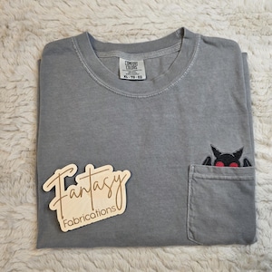 May include: A grey short-sleeved t-shirt with a chest pocket featuring an embroidered black bat with red eyes. The shirt has a Comfort Colors label indicating size XL. A wooden sign with the text Fantasy Fabrications is placed on the shirt.