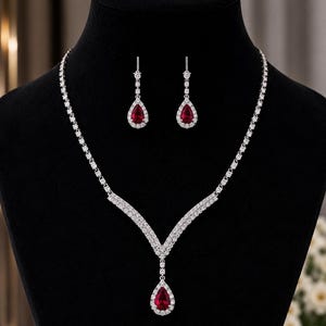Puede incluir: Un conjunto de joyería que consta de un collar y un par de pendientes colgantes. El collar presenta un diseño en forma de V con piedras transparentes y una piedra roja central en forma de pera. Los pendientes a juego tienen una piedra roja.