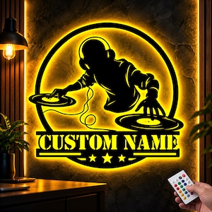 Puede incluir: Un letrero de pared circular que presenta la silueta negra de un DJ con auriculares operando dos tocadiscos, iluminado por una luz LED trasera de color amarillo brillante. El letrero incluye el texto CUSTOM NAME y tres estrellas.