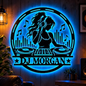 Puede incluir: Un letrero de pared circular que presenta la silueta negra de un DJ con auriculares, colocado detrás de dos tocadiscos y una mesa de mezclas. El diseño incluye notas musicales y un gráfico de ondas sonoras, iluminado por una luz LED azul con el texto DJ MORGAN.