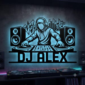May include: A black wall-mounted sign featuring a hooded figure wearing headphones, positioned behind a DJ mixer with two turntables and two speakers. The design includes musical notes and the text DJ ALEX, illuminated by a bright blue backlight.