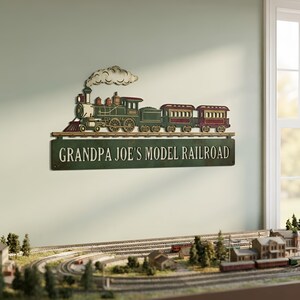 May include: A wall-mounted metal sign featuring a green steam locomotive with two passenger cars and a plume of smoke. Below the train, a rectangular green plaque displays the white text "GRANDPA JOE'S MODEL RAILROAD" in a bold font.