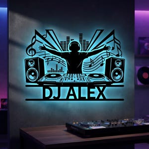 Könnte beinhalten: Ein schwarzes Wand-Schild mit der Silhouette eines DJs mit erhobenen Armen hinter einem Mischpult, flankiert von Lautsprechern und Musiknoten. Das Design ist mit einem hellblauen Licht hinterleuchtet und zeigt den Text DJ ALEX.