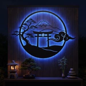 Könnte beinhalten: Ein kreisförmiges Wandkunstobjekt aus schwarzem Metall mit der Silhouette eines Torii-Tors, einer Kiefer und eines Berggipfels, das von hinten durch einen hellblauen LED-Lichtring beleuchtet wird und an einer dunklen Holzplankenwand montiert ist.