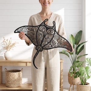 May include: A black laser-cut wall art piece shaped like a manta ray, featuring intricate cut-out patterns of swirling ocean waves, bubbles, and coral reef textures throughout its wings and body, with a long, slender tail extending downward.