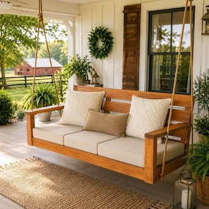 May include: A wooden porch swing suspended by thick ropes, featuring a natural wood frame, three cream-colored seat cushions, two square cream-colored throw pillows with a subtle textured pattern, and one rectangular tan lumbar pillow.