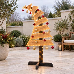 May include: A decorative wooden Christmas tree with a curved top, featuring horizontal light-coloured wood planks attached to a dark vertical post and base. The tree is adorned with warm white string lights and hanging red and gold spherical ornaments.