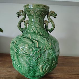 May include: A glossy green ceramic vase featuring a prominent relief of a dragon wrapping around the body. The vessel has a wide neck and two symmetrical handles shaped like dragon heads, with a visible crackle glaze finish throughout the surface.