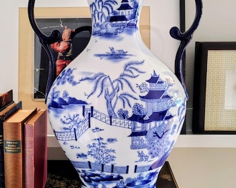 Może przedstawiać: Biały ceramiczny wazon z niebieskim wzorem wierzby, przedstawiający szczegółowe ilustracje pagód, mostów, drzew i łodzi na wodzie. Wazon ma szeroką szyjkę, zaokrąglony korpus i dwa ciemnoniebieskie, zakrzywione uchwyty po obu stronach.