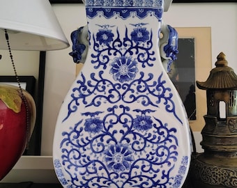 Może przedstawiać: Biały ceramiczny wazon z niebieskim wzorem kwiatowym i misternymi zawijasami. Wazon posiada zdejmowaną pokrywkę zwieńczoną małą rzeźbioną figurką, dwa boczne uchwyty w kształcie głów zwierząt oraz prostokątną podstawę.