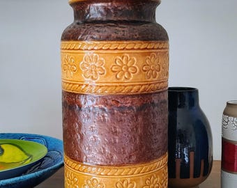 Może przedstawiać: Wysoki cylindryczny wazon ceramiczny z naprzemiennymi pasami ciemnobrązowego, teksturowanego szkliwa i jaśniejszymi sekcjami w kolorze musztardowym. Żółte pasy ozdobione są tłoczonymi wzorami kwiatowymi oraz obwódkami przypominającymi sznur.