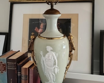 May include: A table lamp featuring a pale green ceramic urn-shaped base with a white relief figure of a person holding a vessel. The lamp has ornate gold-toned metal handles, a matching gold-toned base, and a textured gold-toned drum lampshade.