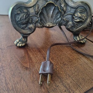 May include: A dark metallic lamp base featuring intricate relief carvings of floral motifs and a central shell design. The base stands on four claw-style feet and includes a brown electrical cord with a two-prong plug extending from the bottom.