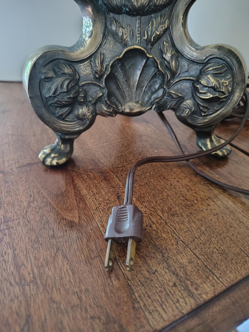 May include: A dark metallic lamp base featuring intricate relief carvings of floral motifs and a central shell design. The base stands on four claw-style feet and includes a brown electrical cord with a two-prong plug extending from the bottom.