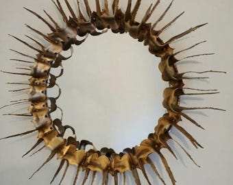 May include: A circular wall mirror featuring a decorative frame composed of numerous thin, pointed, gold-toned metal spikes arranged in a sunburst pattern. The frame has a textured, rippled appearance with varying shades of metallic gold and bronze.