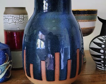 Może przedstawiać: Ceramiczny wazon z błyszczącą, głęboko niebieską glazurą w górnej części i matową podstawą w kolorze terakoty. Wzór charakteryzuje się pionowymi pasami w kolorze terakoty, które biegną w górę od dolnej krawędzi naczynia.