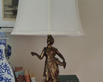 Może przedstawiać: Lampa stołowa z figurą w kolorze brązu przedstawiającą postać w powłóczystych szatach, stojącą na okrągłej podstawie z białego marmuru i ozdobnym metalowym cokole. Lampę wieńczy duży, kremowy abażur z tkaniny z pionowymi szwami.