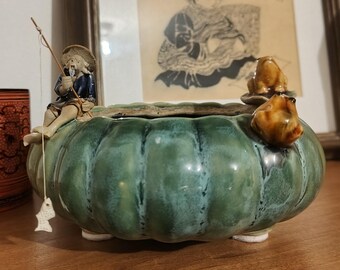 Może przedstawiać: Okrągła, zielona, szkliwiona doniczka ceramiczna o żłobkowanym kształcie przypominającym dynię. Mała figurka w słomkowym kapeluszu i niebieskim ubraniu siedzi na krawędzi, trzymając wędkę z małą białą rybką zwisającą na żyłce.