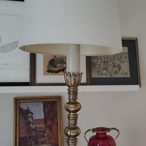 May include: A table lamp with a tall, off-white cylindrical fabric shade and a metallic brass-toned base. The base features a central column with stacked decorative spheres and a wide, ornate, scrolled foot with floral relief detailing.