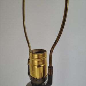 May include: A brass-colored lamp socket assembly featuring a central threaded bulb holder and a tall, U-shaped metal harp attached to the base. The metallic finish shows signs of wear, and the structure is mounted atop a cylindrical white lamp base.