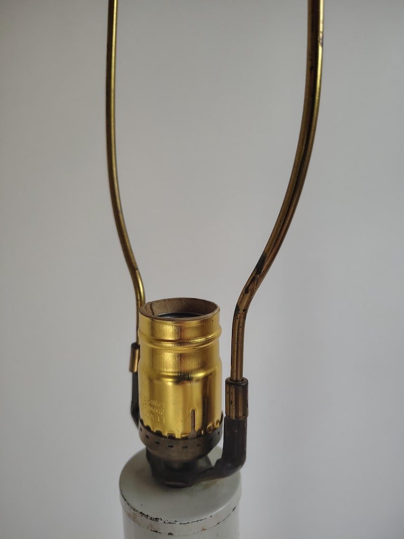 May include: A brass-colored lamp socket assembly featuring a central threaded bulb holder and a tall, U-shaped metal harp attached to the base. The metallic finish shows signs of wear, and the structure is mounted atop a cylindrical white lamp base.