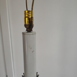 May include: A lamp socket assembly featuring a gold-toned metal light bulb socket mounted atop a cylindrical white column. The base of the column is adorned with a decorative metal leaf-like trim in a muted bronze finish, with a harp attached.