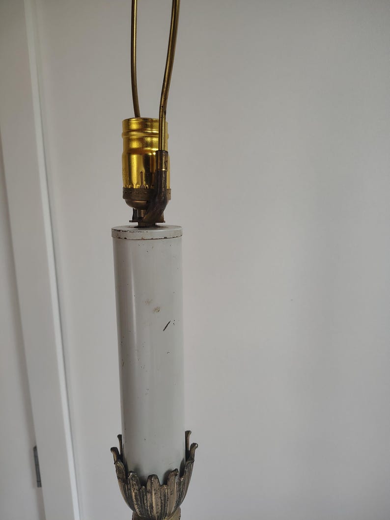 May include: A lamp socket assembly featuring a gold-toned metal light bulb socket mounted atop a cylindrical white column. The base of the column is adorned with a decorative metal leaf-like trim in a muted bronze finish, with a harp attached.