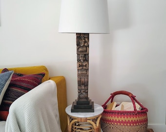 Może przedstawiać: Lampa stołowa z wysoką, prostokątną brązową podstawą z rzeźbionymi figurami i prostym białym abażurem. Lampa stoi na małym, okrągłym stoliku pomocniczym z rattanu z marmurowym blatem obok plecionego kosza wypełnionego książkami i drewnianą skrzynką.