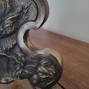 May include: A metallic object with a dark, antiqued bronze finish featuring intricate, raised relief carvings of floral and leaf motifs. The structure has a curved, ornate silhouette with a prominent scroll-like edge and a textured surface.