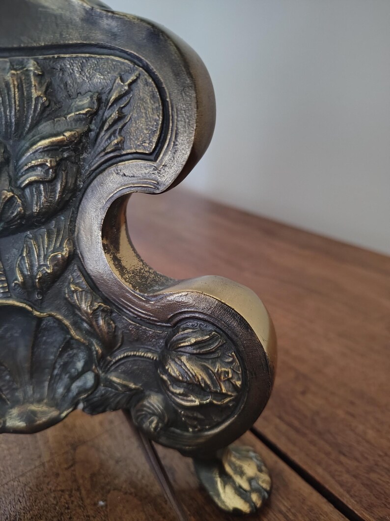 May include: A metallic object with a dark, antiqued bronze finish featuring intricate, raised relief carvings of floral and leaf motifs. The structure has a curved, ornate silhouette with a prominent scroll-like edge and a textured surface.